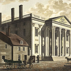 Two-storied Classical style building with columned front entrance stands near smaller buildings along cobblestoned street. Horse-drawn vehicles and pedestrians pass by buildings