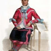 Native American woman wearing a hat, jacket and skirt over a blouse and leggings, her elbow resting on a table.