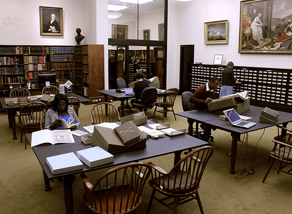 Explore button: Diverse group of researchers working in Library Reading Room with books open on tables.