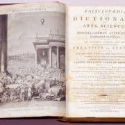 Open book. Crowded outdoor scene and classical-style architecture on left. Title page on right.