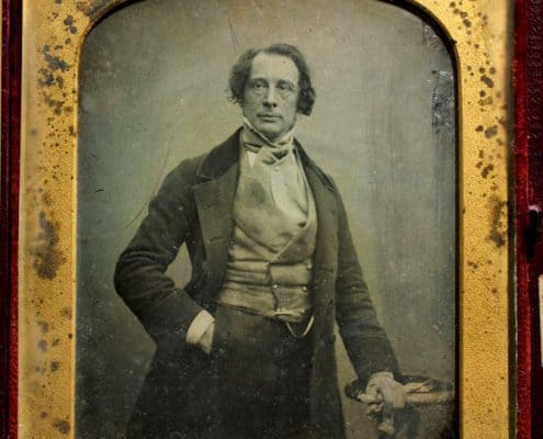 Charles Dickens with right hand in pocket and left hand resting on a small table.