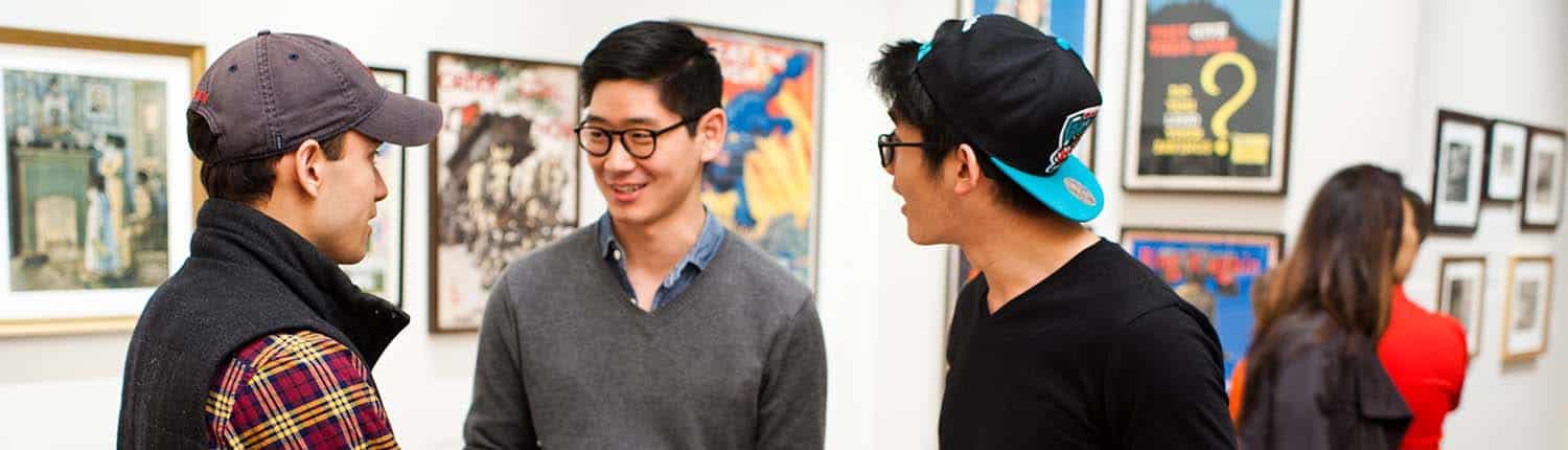 Diverse group of young adults smile and talk together in a Library Company exhibition gallery with framed prints on the wall.
