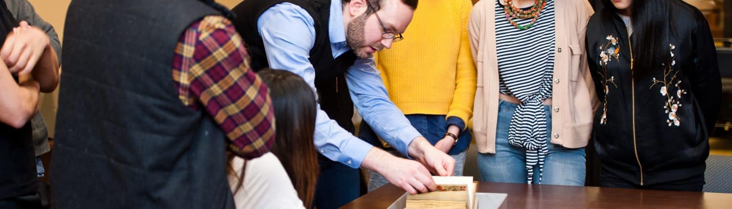 Group of students including white women and Asian American woman peer down at object white man professor presents at Library Company.