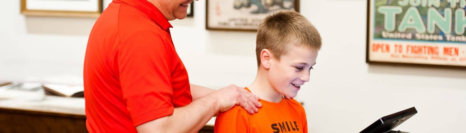 A white man holding shoulders of smiling white boy look down at an iPad in the Library Company exhibition gallery with framed prints on wall.