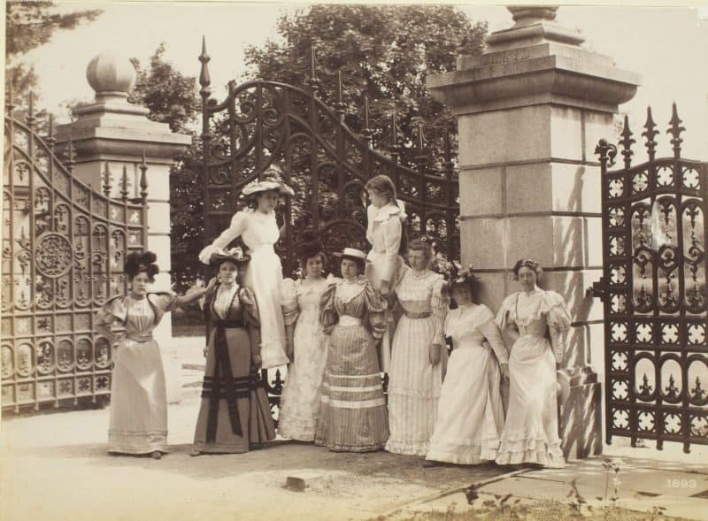 Donate button: Late 19th-century, well-dressed white women and girls standing in front of a large ornate gate.