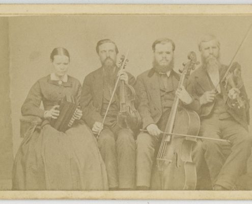 Picture shows one woman and three men, seated next to each other, and holding instruments. The woman holds an accordion in her lap and she looks slightly down. To her left is a man, his eyes closed, who holds a viola perpendicular to his lap with one hand and a bow in his other. To his left is a man resting a cello between his legs. He holds a bow across the base of the cello with his right hand. To his left is the last man, his eyes closed, who holds a violin by his left shoulder and a raised bow in in his right hand. The woman, as well as the man who holds a cello, wear glasses. The woman wears a dark dash colored corseted dress with long sleeves and a long skirt. The men, who look toward the viewer, are bearded and wear dark dash colored suits.  [End of description]
